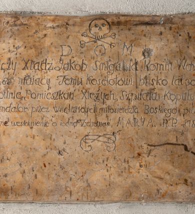 Zdjęcie nr 1: Tablica o kształcie leżącego prostokąta, na której w części górnej wyryto czaszkę powyżej skrzyżowanych piszczeli. Niżej umieszczono inskrypcję „D[eo] O[ptimo] M[aximo] / Tu lezy Xiądz Jakob Smigalski Komm(endarius) Wawrzene(censis) / Lat 85 maiący Temu kościołowi blisko lat 30 służą/cy, pilnie, Pomieszkań Xiężych, Szpitala kopuły, etc / Fundator przez wnętrznosci miłosierdzia Boskiego prosi o po/bożne westchnienie i o iedno Zdrowaś MARYA. R(oku) P(ańskiego) 1789”. Dołem inskrypcję zamyka fragment plecionki.  