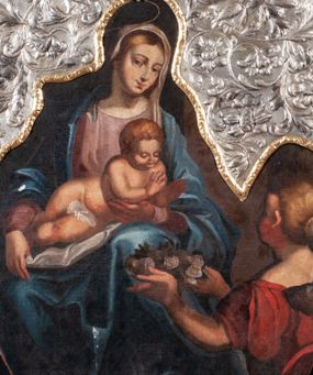Zdjęcie nr 1: Obraz w kształcie owalu malowany na płótnie naklejonym na deskę. W centrum Matka Boska z Dzieciątkiem na kolanach. Maria w pozycji siedzącej, ukazana całopostaciowo, lewą ręką ugiętą w łokciu podtrzymuje Dzieciątko w okolicy klatki piersiowej. Głowa pochylona nieco w lewą stronę. Twarz o rysach młodej kobiety, oczy brązowe, nos długi, usta wąskie. Karnacja jasna, z rumieńcami na policzkach; włosy jasne, przykryte maforionem. Maria jest ubrana w suknię z długimi rękawami w kolorze jasnoróżowym, na którą zarzucony jest obfity, błękitny płaszcz okrywający ramiona. Spod szat widoczna lewa, bosa stopa. Nad głową malowany nimb, powyżej powtórzony w trybowanej blasze; złocony. Dzieciątko jest nagie, leży na kolanach Matki z głową pochyloną lekko do przodu. Ręce  ma ugięte, rozchylone w geście błogosławieństwa. Włosy ma krótkie, gęste, jasnobrązowe, twarz okrągłą z krótkim nosem, wąskimi ustami i półprzymkniętymi powiekami. Z prawej strony, w dole kompozycji, na pierwszym planie, ukazana klęcząca kobieta. W obu wyciągniętych przed siebie rękach trzyma tacę, na której leżą jasnoróżowe róże. Na głowie ma dekoracyjnie upięty kok, w który wpleciony jest pas materiału. Ubrana jest w ciemnoczerwoną suknię z krótkimi rękawami i szerokim dekoltem. Tło kompozycji jednolite, ciemne. Na górną część obrazu nałożony okład ze srebrnej blachy repusowanej w motywy wici roślinno-kwiatowej. Wewnętrzna krawędź okładu obwiedziona pozłoconym pasem blachy, z repusowanym ornamentem roślinnym. Na krawędzi punca kontrybucyjna „CE”.