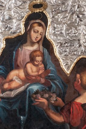 Zdjęcie nr 1: Obraz w kształcie owalu malowany na płótnie naklejonym na deskę. W centrum Matka Boska z Dzieciątkiem na kolanach. Maria w pozycji siedzącej, ukazana całopostaciowo, lewą ręką ugiętą w łokciu podtrzymuje Dzieciątko w okolicy klatki piersiowej. Głowa pochylona nieco w lewą stronę. Twarz o rysach młodej kobiety, oczy brązowe, nos długi, usta wąskie. Karnacja jasna, z rumieńcami na policzkach; włosy jasne, przykryte maforionem. Maria jest ubrana w suknię z długimi rękawami w kolorze jasnoróżowym, na którą zarzucony jest obfity, błękitny płaszcz okrywający ramiona. Spod szat widoczna lewa, bosa stopa. Nad głową malowany nimb, powyżej powtórzony w trybowanej blasze; złocony. Dzieciątko jest nagie, leży na kolanach Matki z głową pochyloną lekko do przodu. Ręce  ma ugięte, rozchylone w geście błogosławieństwa. Włosy ma krótkie, gęste, jasnobrązowe, twarz okrągłą z krótkim nosem, wąskimi ustami i półprzymkniętymi powiekami. Z prawej strony, w dole kompozycji, na pierwszym planie, ukazana klęcząca kobieta. W obu wyciągniętych przed siebie rękach trzyma tacę, na której leżą jasnoróżowe róże. Na głowie ma dekoracyjnie upięty kok, w który wpleciony jest pas materiału. Ubrana jest w ciemnoczerwoną suknię z krótkimi rękawami i szerokim dekoltem. Tło kompozycji jednolite, ciemne. Na górną część obrazu nałożony okład ze srebrnej blachy repusowanej w motywy wici roślinno-kwiatowej. Wewnętrzna krawędź okładu obwiedziona pozłoconym pasem blachy, z repusowanym ornamentem roślinnym. Na krawędzi punca kontrybucyjna „CE”.
