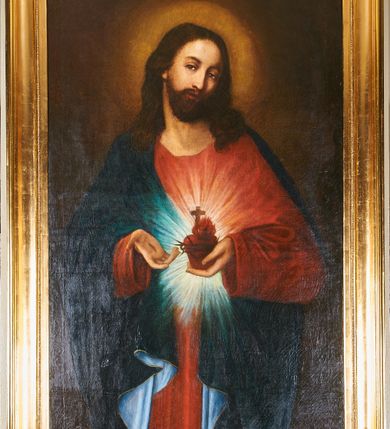 Zdjęcie nr 2: Obraz w kształcie stojącego prostokąta w złoconej, profilowanej ramie. Na nim postać Chrystusa ukazanego w ujęciu do kolan, stojącego centralnie, z głową przechyloną w stronę lewego barku, sercem w lewej dłoni i wskazując na nie prawą. Głowa zwrócona w lewo,  wzrok skierowany na widza nos prosty, usta wąskie; twarz okala bujny, ciemnobrązowy zarost, falowane włosy opadają na ramiona i plecy. Wokół głowy świetlisty nimb. Chrystus jest ubrany w czerwoną tunikę z długimi rękawami i granatowy, podbity bielą płaszcz lamowany złotem, który przerzucony jest przez prawy bark. Trzymane w dłoni serce jest otoczone koroną cierniową, zwieńczone płomieniem i krzyżem łacińskim, otacza je jasna gloria promienista. Tło jednolite, ciemne.