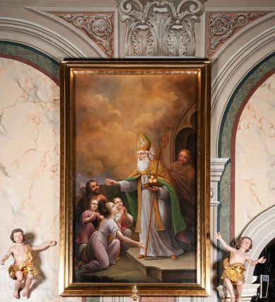 Zdjęcie nr 23: Obraz w kształcie stojącego prostokąta. W centrum, nieco na prawo od osi obrazu, postać św. Mikołaja, który stoi na stopniu przed wejściem do kościoła, odwrócony w trzech czwartych w stronę klęczącej przed nim rodziny. Lewą ręką przytrzymuje pastorał, na dłoni trzyma zamkniętą księgę, na której leżą trzy złote kule, prawą błogosławi klęczących. Ma okrągłą twarz z wydatnymi oczami, prostym nosem oraz grubymi ustami. Siwe włosy spływają mu na plecy, długa broda sięga mostka. Ubrany jest w strój biskupi: albę dekorowaną dołem haftem z motywami roślinnymi oraz stułę, założoną na wierzch kapę spiętą pod szyją i infułę. Trzy ostatnie elementy stroju są zdobione złotą, haftowaną dekoracją wykorzystującą motywy roślinne; końce stuły oraz infuła zdobione krzyżami greckimi. Na piersi widoczny zawieszony na szyi pektorał. Przed świętym znajdują się cztery osoby. Kobieta na pierwszym planie klęczy na prawym kolanie, prawą rękę otwiera do boku, lewą zbliża do piersi, głowę ma uniesioną w kierunku świętego. Ma pogodny wyraz twarzy, duże oczy, prosty nos i jasne włosy spięte w kok. Jest ubrana w różową suknię i żółty płaszcz przewieszony przez lewe ramię. Za nią z prawej strony młoda dziewczyna, która składa ręce w modlitewnym geście spoglądając do góry. Jest ubrana w białą suknię i czerwony płaszcz. Po lewej mała dziewczynka, stoi na lekko ugiętych nogach, składa ręce i z uśmiechem patrzy na świętego, jasne włosy ma rozpuszczone, opadające na plecy. Jest ubrana w białą sukienkę i różową tunikę z szerokim dekoltem. Za nimi klęczy mężczyzna, który również składa ręce, patrzy z zachwytem na postać biskupa; jest ubrany w brązowy strój. Za nimi w tle widać kilka domów i kawałek nieba z księżycem. Za św. Mikołajem, z drzwi kościoła przed którym stoi, wychodzi mężczyzna cały okryty brązowym płaszczem. Górną część kompozycji wypełniają skłębione chmury w odcieniu złocistobrunatnym. Obraz ujęty profilowaną, złoconą ramą. 