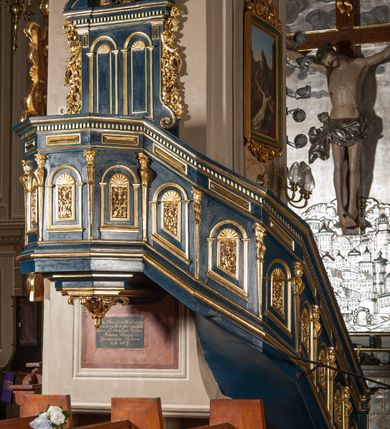 Zdjęcie nr 1: Ambona wisząca, przyścienna, z wejściem od prawej strony w postaci jednobiegowych schodów przylegających do filara, z balustradą. Kosz na planie ośmioboku, z parapetem, od dołu zamknięty uskokowym, zwężającym się w dół profilowaniem z podwieszonym kwiatonem. Ściany kosza i balustrady schodów podzielone na segmenty smukłymi pilastrami hermowymi zdobionymi cekinami, z wolutami. W każdym polu płycina w kształcie pionowego prostokąta, wypełniona ażurowym ornamentem roślinnym z cekinem, zamknięta od góry łukiem półkolistym wypełnionym muszlą. Wokół płycin dodatkowe profilowane obramienia powtarzające ich kształt. Płyciny na balustradzie mają dolną krawędź ustawioną pod skosem tak, by dopasować się do krzywizny wznoszenia (podobnie jest w przypadku innych elementów dekorujących powierzchnię balustrady). Pilastry dzielące balustradę na pola wspierają się na cokole i dźwigają belkowanie, którego fryz wypełniają prostokątne rauty, a gzyms zdobi ząbkowanie. Zaplecek w kształcie pionowego prostokąta dekorowanego dwiema ustawionymi obok siebie płycinami w kształcie wydłużonych prostokątów zamkniętych łukami półkolistymi wypełnionymi muszlami. Wzdłuż bocznych krawędzi zaplecka rodzaj pilastrów wspierających belkowanie dekorowane ząbkowaniem. Po bokach zaplecka dodatkowo uszaki utworzone z ornamentu akantowego i pąków kwiatów. W zwieńczeniu kartusz z monogramem „IHS”w okrągłym polu, z krzyżem zatkniętym na szczycie, flankowany fragmentami przerwanego przyczółka. Struktura malowana na kolor niebieski; profile, ornamenty i detale złocone.