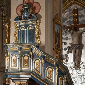 Zdjęcie nr 1: Ambona wisząca, przyścienna, z wejściem od prawej strony w postaci jednobiegowych schodów przylegających do filara, z balustradą. Kosz na planie ośmioboku, z parapetem, od dołu zamknięty uskokowym, zwężającym się w dół profilowaniem z podwieszonym kwiatonem. Ściany kosza i balustrady schodów podzielone na segmenty smukłymi pilastrami hermowymi zdobionymi cekinami, z wolutami. W każdym polu płycina w kształcie pionowego prostokąta, wypełniona ażurowym ornamentem roślinnym z cekinem, zamknięta od góry łukiem półkolistym wypełnionym muszlą. Wokół płycin dodatkowe profilowane obramienia powtarzające ich kształt. Płyciny na balustradzie mają dolną krawędź ustawioną pod skosem tak, by dopasować się do krzywizny wznoszenia (podobnie jest w przypadku innych elementów dekorujących powierzchnię balustrady). Pilastry dzielące balustradę na pola wspierają się na cokole i dźwigają belkowanie, którego fryz wypełniają prostokątne rauty, a gzyms zdobi ząbkowanie. Zaplecek w kształcie pionowego prostokąta dekorowanego dwiema ustawionymi obok siebie płycinami w kształcie wydłużonych prostokątów zamkniętych łukami półkolistymi wypełnionymi muszlami. Wzdłuż bocznych krawędzi zaplecka rodzaj pilastrów wspierających belkowanie dekorowane ząbkowaniem. Po bokach zaplecka dodatkowo uszaki utworzone z ornamentu akantowego i pąków kwiatów. W zwieńczeniu kartusz z monogramem „IHS”w okrągłym polu, z krzyżem zatkniętym na szczycie, flankowany fragmentami przerwanego przyczółka. Struktura malowana na kolor niebieski; profile, ornamenty i detale złocone.