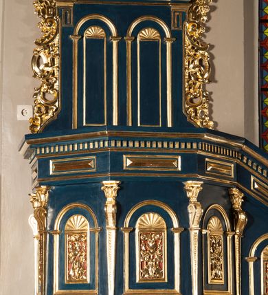 Zdjęcie nr 3: Ambona wisząca, przyścienna, z wejściem od prawej strony w postaci jednobiegowych schodów przylegających do filara, z balustradą. Kosz na planie ośmioboku, z parapetem, od dołu zamknięty uskokowym, zwężającym się w dół profilowaniem z podwieszonym kwiatonem. Ściany kosza i balustrady schodów podzielone na segmenty smukłymi pilastrami hermowymi zdobionymi cekinami, z wolutami. W każdym polu płycina w kształcie pionowego prostokąta, wypełniona ażurowym ornamentem roślinnym z cekinem, zamknięta od góry łukiem półkolistym wypełnionym muszlą. Wokół płycin dodatkowe profilowane obramienia powtarzające ich kształt. Płyciny na balustradzie mają dolną krawędź ustawioną pod skosem tak, by dopasować się do krzywizny wznoszenia (podobnie jest w przypadku innych elementów dekorujących powierzchnię balustrady). Pilastry dzielące balustradę na pola wspierają się na cokole i dźwigają belkowanie, którego fryz wypełniają prostokątne rauty, a gzyms zdobi ząbkowanie. Zaplecek w kształcie pionowego prostokąta dekorowanego dwiema ustawionymi obok siebie płycinami w kształcie wydłużonych prostokątów zamkniętych łukami półkolistymi wypełnionymi muszlami. Wzdłuż bocznych krawędzi zaplecka rodzaj pilastrów wspierających belkowanie dekorowane ząbkowaniem. Po bokach zaplecka dodatkowo uszaki utworzone z ornamentu akantowego i pąków kwiatów. W zwieńczeniu kartusz z monogramem „IHS”w okrągłym polu, z krzyżem zatkniętym na szczycie, flankowany fragmentami przerwanego przyczółka. Struktura malowana na kolor niebieski; profile, ornamenty i detale złocone.