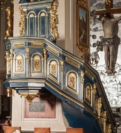 Zdjęcie nr 2: Ambona wisząca, przyścienna, z wejściem od prawej strony w postaci jednobiegowych schodów przylegających do filara, z balustradą. Kosz na planie ośmioboku, z parapetem, od dołu zamknięty uskokowym, zwężającym się w dół profilowaniem z podwieszonym kwiatonem. Ściany kosza i balustrady schodów podzielone na segmenty smukłymi pilastrami hermowymi zdobionymi cekinami, z wolutami. W każdym polu płycina w kształcie pionowego prostokąta, wypełniona ażurowym ornamentem roślinnym z cekinem, zamknięta od góry łukiem półkolistym wypełnionym muszlą. Wokół płycin dodatkowe profilowane obramienia powtarzające ich kształt. Płyciny na balustradzie mają dolną krawędź ustawioną pod skosem tak, by dopasować się do krzywizny wznoszenia (podobnie jest w przypadku innych elementów dekorujących powierzchnię balustrady). Pilastry dzielące balustradę na pola wspierają się na cokole i dźwigają belkowanie, którego fryz wypełniają prostokątne rauty, a gzyms zdobi ząbkowanie. Zaplecek w kształcie pionowego prostokąta dekorowanego dwiema ustawionymi obok siebie płycinami w kształcie wydłużonych prostokątów zamkniętych łukami półkolistymi wypełnionymi muszlami. Wzdłuż bocznych krawędzi zaplecka rodzaj pilastrów wspierających belkowanie dekorowane ząbkowaniem. Po bokach zaplecka dodatkowo uszaki utworzone z ornamentu akantowego i pąków kwiatów. W zwieńczeniu kartusz z monogramem „IHS”w okrągłym polu, z krzyżem zatkniętym na szczycie, flankowany fragmentami przerwanego przyczółka. Struktura malowana na kolor niebieski; profile, ornamenty i detale złocone.