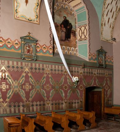 Zdjęcie nr 2: Prezbiterium: 
Sklepienie kolebkowe z lunetami; trójprzęsłowe. Pola malowane  są na kolor biały z centralnie umieszczonymi kolistymi medalionami. W przęśle pierwszym od ołtarza znajduje się motyw krzyża greckiego na tle promienistej glorii; w drugim gołębica Ducha Świętego na tle analogicznej glorii zamknięta w polu przypominającym sześciolistny kwiat o błękitno-złotych płatkach; w trzecim znak Trójcy Świętej w formie trzech przecinających się okręgów z napisem „TRI/NI/TAS”, otoczonych promieniami i płomienistymi półokręgami. Pozostałe partie pól sklepienia wypełnione są złoconymi językami ognia. Wokół pól sklepienia biegnie ornament geometryczny w kolorze turkusowym z elementami ugru i ultramaryny. Lunety malowane są na kolor jasnoniebieski. W każdej z nich znajdują się medaliony o kształcie zbliżonym do czwórliścia, wewnątrz których, od wschodu jest: słońce, planeta, gwiazda, księżyc, monogram Chrystusa, napis „CA/RI/TAS”.  Ściany boczne w około połowie wysokości podzielone na dwa poziomy. Dolny wypełniony jest geometryczną dekoracją w formie fryzu ze złoconych rombów, naprzemiennie większych i mniejszych. Wewnątrz figur równoramienne krzyże oraz motyw uproszczonych liści. Fryz ujęty od góry i dołu abstrakcyjną dekoracją o charakterze geometrycznym. Tła malowane są na kolor ciemnoczerwony oraz zielony. Poziom górny malowany jest na bladoróżowo. Na wysokości parapetów okiennych fryz ze stylizowanych motywów kwiatowych w kolorze żółtym, zielonym i brązowym. W ościeżach dekoracja ramowa w kolorze turkusowym i bladożółtym wzbogacona o malowane ciemniejszą farbą motywy geometryczne. Okna ściany południowej zastąpione blendami; w nich motyw karpiej łuski wypełnionej palmetą.

Łuk arkady tęczowej:
Na licu arkady tęczowej znajduje się figuralne przedstawienie stygmatyzacji św. Franciszka. Z lewej strony św. Franciszek, zwrócony w lewą stronę, z rozłożonymi szeroko rękami i prawą nogą cofniętą do tyłu. Ubrany w brązowy habit franciszkański. Z prawej strony Chrystus, ukazany naturalnej wielkości, jako cherubin o dużych, czerwonych skrzydłach. Z jego ran wychodzą promienie tworzące stygmaty na ciele świętego. Scena rozgrywa się na kamienistej drodze, na tle płomieni. Kompozycja ujęta otokiem z białych chmur. Na podłuczu arkady symetryczna dekoracja geometryczna w formie motywu „jodełki” przedzielonego wstęgą. W osi ośmioboczne pole ze znakiem krzyża greckiego z napisem „Cristus/Jesus”. Kolorystyka utrzymana w ciemnej zieleni, ceglastej czerwieni oraz w kolorze złotym.

Nawa główna:
Sklepienie kolebkowe z lunetami, czteroprzęsłowe. Pola malowane kolorem jasnoniebieskim. Cała długość nawy połączona dekoracyjnym pasem w kolorach turkusowym, szafirowym oraz żółtym. W każdym z przęseł pośrodku czteorolistne  pole otoczone glorią promienistą zawierające znaki, od wschodu: Alfa i Omega, monogram maryjny, kielich z hostią, IHS. Lunety malowane na jasnożółto, pośrodku każdej owoidalne pole w kolorze szafirowym: od wschodu i zachodu po dwóch stronach korona i napis „VITA / AETERNA”, od południa waga i napis „IUSTI/TIA”, dalej miecz i napis „FORTI\TUDO”, od północy beczka i napis „TEM/PE/RANTIA, wąż i napis „PRUDENTIA”. Obramienia lunet z motywem abstrakcyjnym. Pasy gurtów z ludowymi motywami kwiatowymi i kłosami zboża na żółtym tle. Ściany boczne w partii cokołu malowane są na kolor różowy, powyżej aż do linii parapetów okiennych: miodowo-żółty. Na obramieniach arkadowych nisz jest stylizowana dekoracja roślinna, w podłuczach i ościeżach arkad dekoracja analogiczna do tej z podłucza arkady tęczy. Na ścianach pomiędzy niszami godła franciszkańskie zamknięte w okrągłych medalionach. Na linii parapetów geometryczny fryz z przecinających się arkadek uzupełnionych o motywy roślinne. W rozglifieniach okiennych naprzemiennie motywy geometryczne i roślinne w kolorach błękitnym, pomarańczoym i dwóch odcieniach żółci. Arkada chóru muzycznego o podziale ramowym w kolorach jasnozielonym, pomarańczowym i żółtym.