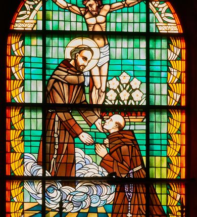 Zdjęcie nr 1: Witraż w kształcie stojącego prostokąta zamkniętego łukiem pełnym . W centrum rozgrywająca się we wnetrzu scena, w której św. Franciszek na obłokach ukazuje się klęczącemu bratu Kosibie. Po lewej stronie stoi św. Franciszek wyciągający ręce w stronę brata Alojzego. Spogląda na niego, ma ciemne oczy, prosty nos, wąskie usta, ciemnobrązową brodę i takiego koloru włosy z wycięta tonsurą; głowę otacza nimb. Ubrany jest w habit przewiązany w pasie sznurem. Brak Kosiba klęczy bliżej widza, został ukazany z profilu, ręce wznosi w stronę św. Franciszka. Twarz o cechach portretowych, oczy ciemne, nos prosty, usta wąskie, głowa niemal łysa. Jest brany w habit franciszkański przepasany sznurkiem, z przywiązanym różańcem. W tle krucyfiks oraz kosz z kwitnącymi kwiatami lilii zawieszone na zielonej ścianie. Scenę otacza bordiura wypełniona w górnej części wspartym na głowicach łukiem z przecinających się półkoli, półokrągłymi, żółtymi elementami na tle zieleni po bokach oraz rozetami w dolnej partii. Między nimi napis „W 50-TĄ ROCZNICĘ NADZIN DLA NIEBA / BRATA ALOJZEGO-KOSIBY 1989”. Kolorystyka ciepła z dominacją brązów i żółci oraz zieleni.