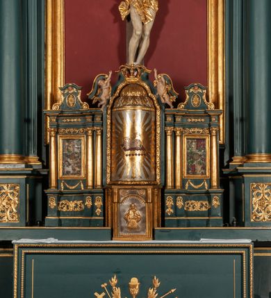 Zdjęcie nr 5: Ołtarz drewniany, dwustronny, obecnie przyścienny, architektoniczny, jednokondygnacyjny ze zwieńczeniem, trójosiowy, z bramkami. Mensa kamienna, prostopadłościenna z antepedium ujętym w profilowaną i złoconą ramę, dekorowaną kimationem jońskim i kampanulami. W polu płaskorzeźbione przedstawienie kielicha z hostią ujętego dwoma otwartymi księgami z napisem na prawej „ECCE / PANIS / ANGE/LORUM”, na lewej „EVAN/GE/LIUM”; liśćmi laurowymi, arma Christi; u podstawy kielicha nałożona korona cierniowa. W dolnych narożach antepedium dekoracja z miękko zawijającej się wstęgi regencyjnej. Tabernakulum o rozbudowanych architektonicznych formach, jednokondygnacyjne, trójosiowe, każda z osi flankowana parą kolumienek i zakończona zwieńczeniem. W osi środkowej tabernakulum metalowe: w dolnej części w formie prostopadłościennej skrzynki z wizerunkiem Chrystusa, w górnej części obrotowe z przedstawieniem Baranka Bożego, zamknięte ozdobnym zwieńczeniem z parą uskrzydlonych putt i koroną w typie zamkniętym. W polach osi bocznych umieszczone za szybką relikwie świętych ujęte prostokątną, złoconą ramą. Retabulum ustawione na wysokim, dwudzielnym cokole, z bramkami o otworach drzwiowych zamkniętych łukiem kotarowym ściętym, nadproża w formie profilowanego gryzmsu. Pole ołtarza, zarówno osi głównej jak i bocznych ujęte jest kolumnami o gładkich trzonach i złoconych kapitalach kompozytowych ustawionych na tle zwieloktrotnionych pilastrów o kanelowanych trzonach. Podpory dźwigają wyłamane na ich osi, pełne belkowanie z wydatnym, silnie profilowanych gzymsem. Na nim fragmenty przyczółka w formie wolut. Pole osi środkowej ze złoconą ramą zamkniętą łukiem wklęsło-wypukłym z uskokiem. W zwieńczeniu ramy wolutowy kartusz o nieregularnym kształcie, z napisem „CONSUMMATUM / EST”. W centrum pola ołtarzowego drewniany krycyfiks. W osi prawej figura św. Kazimierza Królewicza, w osi lewej figura św. Jadwigi Śląskiej. Rzeźby ustawione w niszach, zamkniętych łukiem wklęsło-wypukłym, na rozbudowanych, wolutowych konsolach dekorowanych z przodu kartuszem utworzonym z akantowego wieńca wypełnionego kratką regencyjną; ponad nim girlanda złożona z kampanul i kwiatów. W niszach ponad figurami umieszczone dużych rozmiarów, złocone muszle palmetowe, oraz nieco niżej dwie gałązki akantu. Ponad niszami rozbudowana płaskorzeźbiona dekoracja w formie struktury utworzonej z wolut o karbowanych wypełnieniach, w jej centrum uskrzydlone główki aniołków. Zwieńczenie w formie wklęsło-wypukłej, zamknięte gzymsem w kształcie łuku nadwieszonego, po bokach flankowane dwoma wazonami z kwiatami. W polu obraz św. Michała Archanioła. Kolorystyka ciemnozielona, ze złoceniami w partiach ornamentów oraz w załamaniach profili architektonicznych. Partie cokołu dekorowane płycinami wypełnionymi ornamentem regencyjnym. Według dokumentacji konserwatorskiej na odwrociu ołtarza znajduje się malowana iluzjonistyczna dekoracja przedstawiająca barokowy ołtarz, postacie Matki Boskiej, św. Jana Ewangelisty oraz świętych franciszkańskich.

