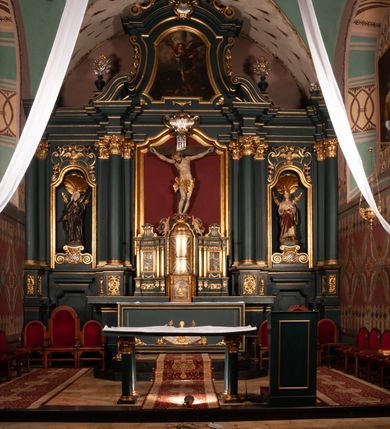 Zdjęcie nr 2: Ołtarz drewniany, dwustronny, obecnie przyścienny, architektoniczny, jednokondygnacyjny ze zwieńczeniem, trójosiowy, z bramkami. Mensa kamienna, prostopadłościenna z antepedium ujętym w profilowaną i złoconą ramę, dekorowaną kimationem jońskim i kampanulami. W polu płaskorzeźbione przedstawienie kielicha z hostią ujętego dwoma otwartymi księgami z napisem na prawej „ECCE / PANIS / ANGE/LORUM”, na lewej „EVAN/GE/LIUM”; liśćmi laurowymi, arma Christi; u podstawy kielicha nałożona korona cierniowa. W dolnych narożach antepedium dekoracja z miękko zawijającej się wstęgi regencyjnej. Tabernakulum o rozbudowanych architektonicznych formach, jednokondygnacyjne, trójosiowe, każda z osi flankowana parą kolumienek i zakończona zwieńczeniem. W osi środkowej tabernakulum metalowe: w dolnej części w formie prostopadłościennej skrzynki z wizerunkiem Chrystusa, w górnej części obrotowe z przedstawieniem Baranka Bożego, zamknięte ozdobnym zwieńczeniem z parą uskrzydlonych putt i koroną w typie zamkniętym. W polach osi bocznych umieszczone za szybką relikwie świętych ujęte prostokątną, złoconą ramą. Retabulum ustawione na wysokim, dwudzielnym cokole, z bramkami o otworach drzwiowych zamkniętych łukiem kotarowym ściętym, nadproża w formie profilowanego gryzmsu. Pole ołtarza, zarówno osi głównej jak i bocznych ujęte jest kolumnami o gładkich trzonach i złoconych kapitalach kompozytowych ustawionych na tle zwieloktrotnionych pilastrów o kanelowanych trzonach. Podpory dźwigają wyłamane na ich osi, pełne belkowanie z wydatnym, silnie profilowanych gzymsem. Na nim fragmenty przyczółka w formie wolut. Pole osi środkowej ze złoconą ramą zamkniętą łukiem wklęsło-wypukłym z uskokiem. W zwieńczeniu ramy wolutowy kartusz o nieregularnym kształcie, z napisem „CONSUMMATUM / EST”. W centrum pola ołtarzowego drewniany krycyfiks. W osi prawej figura św. Kazimierza Królewicza, w osi lewej figura św. Jadwigi Śląskiej. Rzeźby ustawione w niszach, zamkniętych łukiem wklęsło-wypukłym, na rozbudowanych, wolutowych konsolach dekorowanych z przodu kartuszem utworzonym z akantowego wieńca wypełnionego kratką regencyjną; ponad nim girlanda złożona z kampanul i kwiatów. W niszach ponad figurami umieszczone dużych rozmiarów, złocone muszle palmetowe, oraz nieco niżej dwie gałązki akantu. Ponad niszami rozbudowana płaskorzeźbiona dekoracja w formie struktury utworzonej z wolut o karbowanych wypełnieniach, w jej centrum uskrzydlone główki aniołków. Zwieńczenie w formie wklęsło-wypukłej, zamknięte gzymsem w kształcie łuku nadwieszonego, po bokach flankowane dwoma wazonami z kwiatami. W polu obraz św. Michała Archanioła. Kolorystyka ciemnozielona, ze złoceniami w partiach ornamentów oraz w załamaniach profili architektonicznych. Partie cokołu dekorowane płycinami wypełnionymi ornamentem regencyjnym. Według dokumentacji konserwatorskiej na odwrociu ołtarza znajduje się malowana iluzjonistyczna dekoracja przedstawiająca barokowy ołtarz, postacie Matki Boskiej, św. Jana Ewangelisty oraz świętych franciszkańskich.

