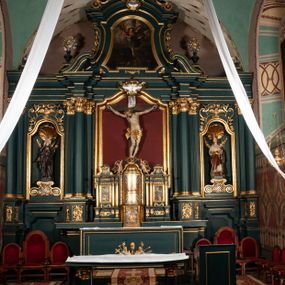 Zdjęcie nr 1: Ołtarz drewniany, dwustronny, obecnie przyścienny, architektoniczny, jednokondygnacyjny ze zwieńczeniem, trójosiowy, z bramkami. Mensa kamienna, prostopadłościenna z antepedium ujętym w profilowaną i złoconą ramę, dekorowaną kimationem jońskim i kampanulami. W polu płaskorzeźbione przedstawienie kielicha z hostią ujętego dwoma otwartymi księgami z napisem na prawej „ECCE / PANIS / ANGE/LORUM”, na lewej „EVAN/GE/LIUM”; liśćmi laurowymi, arma Christi; u podstawy kielicha nałożona korona cierniowa. W dolnych narożach antepedium dekoracja z miękko zawijającej się wstęgi regencyjnej. Tabernakulum o rozbudowanych architektonicznych formach, jednokondygnacyjne, trójosiowe, każda z osi flankowana parą kolumienek i zakończona zwieńczeniem. W osi środkowej tabernakulum metalowe: w dolnej części w formie prostopadłościennej skrzynki z wizerunkiem Chrystusa, w górnej części obrotowe z przedstawieniem Baranka Bożego, zamknięte ozdobnym zwieńczeniem z parą uskrzydlonych putt i koroną w typie zamkniętym. W polach osi bocznych umieszczone za szybką relikwie świętych ujęte prostokątną, złoconą ramą. Retabulum ustawione na wysokim, dwudzielnym cokole, z bramkami o otworach drzwiowych zamkniętych łukiem kotarowym ściętym, nadproża w formie profilowanego gryzmsu. Pole ołtarza, zarówno osi głównej jak i bocznych ujęte jest kolumnami o gładkich trzonach i złoconych kapitalach kompozytowych ustawionych na tle zwieloktrotnionych pilastrów o kanelowanych trzonach. Podpory dźwigają wyłamane na ich osi, pełne belkowanie z wydatnym, silnie profilowanych gzymsem. Na nim fragmenty przyczółka w formie wolut. Pole osi środkowej ze złoconą ramą zamkniętą łukiem wklęsło-wypukłym z uskokiem. W zwieńczeniu ramy wolutowy kartusz o nieregularnym kształcie, z napisem „CONSUMMATUM / EST”. W centrum pola ołtarzowego drewniany krycyfiks. W osi prawej figura św. Kazimierza Królewicza, w osi lewej figura św. Jadwigi Śląskiej. Rzeźby ustawione w niszach, zamkniętych łukiem wklęsło-wypukłym, na rozbudowanych, wolutowych konsolach dekorowanych z przodu kartuszem utworzonym z akantowego wieńca wypełnionego kratką regencyjną; ponad nim girlanda złożona z kampanul i kwiatów. W niszach ponad figurami umieszczone dużych rozmiarów, złocone muszle palmetowe, oraz nieco niżej dwie gałązki akantu. Ponad niszami rozbudowana płaskorzeźbiona dekoracja w formie struktury utworzonej z wolut o karbowanych wypełnieniach, w jej centrum uskrzydlone główki aniołków. Zwieńczenie w formie wklęsło-wypukłej, zamknięte gzymsem w kształcie łuku nadwieszonego, po bokach flankowane dwoma wazonami z kwiatami. W polu obraz św. Michała Archanioła. Kolorystyka ciemnozielona, ze złoceniami w partiach ornamentów oraz w załamaniach profili architektonicznych. Partie cokołu dekorowane płycinami wypełnionymi ornamentem regencyjnym. Według dokumentacji konserwatorskiej na odwrociu ołtarza znajduje się malowana iluzjonistyczna dekoracja przedstawiająca barokowy ołtarz, postacie Matki Boskiej, św. Jana Ewangelisty oraz świętych franciszkańskich.

