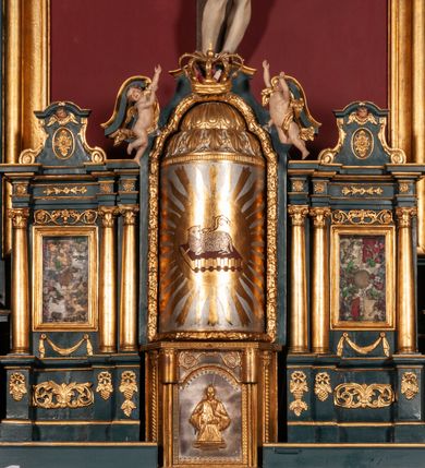 Zdjęcie nr 3: Ołtarz drewniany, dwustronny, obecnie przyścienny, architektoniczny, jednokondygnacyjny ze zwieńczeniem, trójosiowy, z bramkami. Mensa kamienna, prostopadłościenna z antepedium ujętym w profilowaną i złoconą ramę, dekorowaną kimationem jońskim i kampanulami. W polu płaskorzeźbione przedstawienie kielicha z hostią ujętego dwoma otwartymi księgami z napisem na prawej „ECCE / PANIS / ANGE/LORUM”, na lewej „EVAN/GE/LIUM”; liśćmi laurowymi, arma Christi; u podstawy kielicha nałożona korona cierniowa. W dolnych narożach antepedium dekoracja z miękko zawijającej się wstęgi regencyjnej. Tabernakulum o rozbudowanych architektonicznych formach, jednokondygnacyjne, trójosiowe, każda z osi flankowana parą kolumienek i zakończona zwieńczeniem. W osi środkowej tabernakulum metalowe: w dolnej części w formie prostopadłościennej skrzynki z wizerunkiem Chrystusa, w górnej części obrotowe z przedstawieniem Baranka Bożego, zamknięte ozdobnym zwieńczeniem z parą uskrzydlonych putt i koroną w typie zamkniętym. W polach osi bocznych umieszczone za szybką relikwie świętych ujęte prostokątną, złoconą ramą. Retabulum ustawione na wysokim, dwudzielnym cokole, z bramkami o otworach drzwiowych zamkniętych łukiem kotarowym ściętym, nadproża w formie profilowanego gryzmsu. Pole ołtarza, zarówno osi głównej jak i bocznych ujęte jest kolumnami o gładkich trzonach i złoconych kapitalach kompozytowych ustawionych na tle zwieloktrotnionych pilastrów o kanelowanych trzonach. Podpory dźwigają wyłamane na ich osi, pełne belkowanie z wydatnym, silnie profilowanych gzymsem. Na nim fragmenty przyczółka w formie wolut. Pole osi środkowej ze złoconą ramą zamkniętą łukiem wklęsło-wypukłym z uskokiem. W zwieńczeniu ramy wolutowy kartusz o nieregularnym kształcie, z napisem „CONSUMMATUM / EST”. W centrum pola ołtarzowego drewniany krycyfiks. W osi prawej figura św. Kazimierza Królewicza, w osi lewej figura św. Jadwigi Śląskiej. Rzeźby ustawione w niszach, zamkniętych łukiem wklęsło-wypukłym, na rozbudowanych, wolutowych konsolach dekorowanych z przodu kartuszem utworzonym z akantowego wieńca wypełnionego kratką regencyjną; ponad nim girlanda złożona z kampanul i kwiatów. W niszach ponad figurami umieszczone dużych rozmiarów, złocone muszle palmetowe, oraz nieco niżej dwie gałązki akantu. Ponad niszami rozbudowana płaskorzeźbiona dekoracja w formie struktury utworzonej z wolut o karbowanych wypełnieniach, w jej centrum uskrzydlone główki aniołków. Zwieńczenie w formie wklęsło-wypukłej, zamknięte gzymsem w kształcie łuku nadwieszonego, po bokach flankowane dwoma wazonami z kwiatami. W polu obraz św. Michała Archanioła. Kolorystyka ciemnozielona, ze złoceniami w partiach ornamentów oraz w załamaniach profili architektonicznych. Partie cokołu dekorowane płycinami wypełnionymi ornamentem regencyjnym. Według dokumentacji konserwatorskiej na odwrociu ołtarza znajduje się malowana iluzjonistyczna dekoracja przedstawiająca barokowy ołtarz, postacie Matki Boskiej, św. Jana Ewangelisty oraz świętych franciszkańskich.

