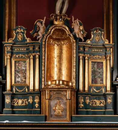 Zdjęcie nr 6: Ołtarz drewniany, dwustronny, obecnie przyścienny, architektoniczny, jednokondygnacyjny ze zwieńczeniem, trójosiowy, z bramkami. Mensa kamienna, prostopadłościenna z antepedium ujętym w profilowaną i złoconą ramę, dekorowaną kimationem jońskim i kampanulami. W polu płaskorzeźbione przedstawienie kielicha z hostią ujętego dwoma otwartymi księgami z napisem na prawej „ECCE / PANIS / ANGE/LORUM”, na lewej „EVAN/GE/LIUM”; liśćmi laurowymi, arma Christi; u podstawy kielicha nałożona korona cierniowa. W dolnych narożach antepedium dekoracja z miękko zawijającej się wstęgi regencyjnej. Tabernakulum o rozbudowanych architektonicznych formach, jednokondygnacyjne, trójosiowe, każda z osi flankowana parą kolumienek i zakończona zwieńczeniem. W osi środkowej tabernakulum metalowe: w dolnej części w formie prostopadłościennej skrzynki z wizerunkiem Chrystusa, w górnej części obrotowe z przedstawieniem Baranka Bożego, zamknięte ozdobnym zwieńczeniem z parą uskrzydlonych putt i koroną w typie zamkniętym. W polach osi bocznych umieszczone za szybką relikwie świętych ujęte prostokątną, złoconą ramą. Retabulum ustawione na wysokim, dwudzielnym cokole, z bramkami o otworach drzwiowych zamkniętych łukiem kotarowym ściętym, nadproża w formie profilowanego gryzmsu. Pole ołtarza, zarówno osi głównej jak i bocznych ujęte jest kolumnami o gładkich trzonach i złoconych kapitalach kompozytowych ustawionych na tle zwieloktrotnionych pilastrów o kanelowanych trzonach. Podpory dźwigają wyłamane na ich osi, pełne belkowanie z wydatnym, silnie profilowanych gzymsem. Na nim fragmenty przyczółka w formie wolut. Pole osi środkowej ze złoconą ramą zamkniętą łukiem wklęsło-wypukłym z uskokiem. W zwieńczeniu ramy wolutowy kartusz o nieregularnym kształcie, z napisem „CONSUMMATUM / EST”. W centrum pola ołtarzowego drewniany krycyfiks. W osi prawej figura św. Kazimierza Królewicza, w osi lewej figura św. Jadwigi Śląskiej. Rzeźby ustawione w niszach, zamkniętych łukiem wklęsło-wypukłym, na rozbudowanych, wolutowych konsolach dekorowanych z przodu kartuszem utworzonym z akantowego wieńca wypełnionego kratką regencyjną; ponad nim girlanda złożona z kampanul i kwiatów. W niszach ponad figurami umieszczone dużych rozmiarów, złocone muszle palmetowe, oraz nieco niżej dwie gałązki akantu. Ponad niszami rozbudowana płaskorzeźbiona dekoracja w formie struktury utworzonej z wolut o karbowanych wypełnieniach, w jej centrum uskrzydlone główki aniołków. Zwieńczenie w formie wklęsło-wypukłej, zamknięte gzymsem w kształcie łuku nadwieszonego, po bokach flankowane dwoma wazonami z kwiatami. W polu obraz św. Michała Archanioła. Kolorystyka ciemnozielona, ze złoceniami w partiach ornamentów oraz w załamaniach profili architektonicznych. Partie cokołu dekorowane płycinami wypełnionymi ornamentem regencyjnym. Według dokumentacji konserwatorskiej na odwrociu ołtarza znajduje się malowana iluzjonistyczna dekoracja przedstawiająca barokowy ołtarz, postacie Matki Boskiej, św. Jana Ewangelisty oraz świętych franciszkańskich.

