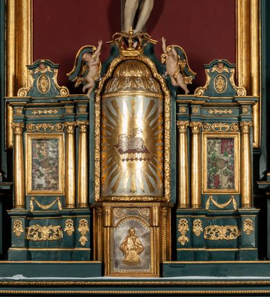 Zdjęcie nr 7: Ołtarz drewniany, dwustronny, obecnie przyścienny, architektoniczny, jednokondygnacyjny ze zwieńczeniem, trójosiowy, z bramkami. Mensa kamienna, prostopadłościenna z antepedium ujętym w profilowaną i złoconą ramę, dekorowaną kimationem jońskim i kampanulami. W polu płaskorzeźbione przedstawienie kielicha z hostią ujętego dwoma otwartymi księgami z napisem na prawej „ECCE / PANIS / ANGE/LORUM”, na lewej „EVAN/GE/LIUM”; liśćmi laurowymi, arma Christi; u podstawy kielicha nałożona korona cierniowa. W dolnych narożach antepedium dekoracja z miękko zawijającej się wstęgi regencyjnej. Tabernakulum o rozbudowanych architektonicznych formach, jednokondygnacyjne, trójosiowe, każda z osi flankowana parą kolumienek i zakończona zwieńczeniem. W osi środkowej tabernakulum metalowe: w dolnej części w formie prostopadłościennej skrzynki z wizerunkiem Chrystusa, w górnej części obrotowe z przedstawieniem Baranka Bożego, zamknięte ozdobnym zwieńczeniem z parą uskrzydlonych putt i koroną w typie zamkniętym. W polach osi bocznych umieszczone za szybką relikwie świętych ujęte prostokątną, złoconą ramą. Retabulum ustawione na wysokim, dwudzielnym cokole, z bramkami o otworach drzwiowych zamkniętych łukiem kotarowym ściętym, nadproża w formie profilowanego gryzmsu. Pole ołtarza, zarówno osi głównej jak i bocznych ujęte jest kolumnami o gładkich trzonach i złoconych kapitalach kompozytowych ustawionych na tle zwieloktrotnionych pilastrów o kanelowanych trzonach. Podpory dźwigają wyłamane na ich osi, pełne belkowanie z wydatnym, silnie profilowanych gzymsem. Na nim fragmenty przyczółka w formie wolut. Pole osi środkowej ze złoconą ramą zamkniętą łukiem wklęsło-wypukłym z uskokiem. W zwieńczeniu ramy wolutowy kartusz o nieregularnym kształcie, z napisem „CONSUMMATUM / EST”. W centrum pola ołtarzowego drewniany krycyfiks. W osi prawej figura św. Kazimierza Królewicza, w osi lewej figura św. Jadwigi Śląskiej. Rzeźby ustawione w niszach, zamkniętych łukiem wklęsło-wypukłym, na rozbudowanych, wolutowych konsolach dekorowanych z przodu kartuszem utworzonym z akantowego wieńca wypełnionego kratką regencyjną; ponad nim girlanda złożona z kampanul i kwiatów. W niszach ponad figurami umieszczone dużych rozmiarów, złocone muszle palmetowe, oraz nieco niżej dwie gałązki akantu. Ponad niszami rozbudowana płaskorzeźbiona dekoracja w formie struktury utworzonej z wolut o karbowanych wypełnieniach, w jej centrum uskrzydlone główki aniołków. Zwieńczenie w formie wklęsło-wypukłej, zamknięte gzymsem w kształcie łuku nadwieszonego, po bokach flankowane dwoma wazonami z kwiatami. W polu obraz św. Michała Archanioła. Kolorystyka ciemnozielona, ze złoceniami w partiach ornamentów oraz w załamaniach profili architektonicznych. Partie cokołu dekorowane płycinami wypełnionymi ornamentem regencyjnym. Według dokumentacji konserwatorskiej na odwrociu ołtarza znajduje się malowana iluzjonistyczna dekoracja przedstawiająca barokowy ołtarz, postacie Matki Boskiej, św. Jana Ewangelisty oraz świętych franciszkańskich.

