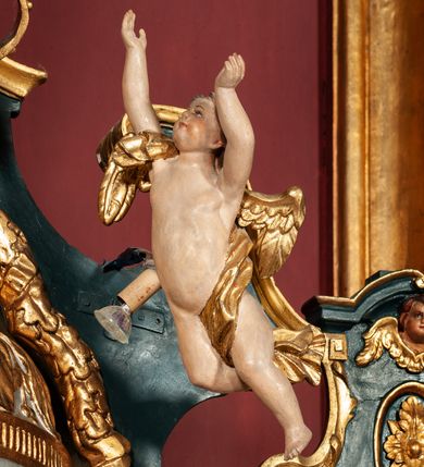 Zdjęcie nr 50: Ołtarz drewniany, dwustronny, obecnie przyścienny, architektoniczny, jednokondygnacyjny ze zwieńczeniem, trójosiowy, z bramkami. Mensa kamienna, prostopadłościenna z antepedium ujętym w profilowaną i złoconą ramę, dekorowaną kimationem jońskim i kampanulami. W polu płaskorzeźbione przedstawienie kielicha z hostią ujętego dwoma otwartymi księgami z napisem na prawej „ECCE / PANIS / ANGE/LORUM”, na lewej „EVAN/GE/LIUM”; liśćmi laurowymi, arma Christi; u podstawy kielicha nałożona korona cierniowa. W dolnych narożach antepedium dekoracja z miękko zawijającej się wstęgi regencyjnej. Tabernakulum o rozbudowanych architektonicznych formach, jednokondygnacyjne, trójosiowe, każda z osi flankowana parą kolumienek i zakończona zwieńczeniem. W osi środkowej tabernakulum metalowe: w dolnej części w formie prostopadłościennej skrzynki z wizerunkiem Chrystusa, w górnej części obrotowe z przedstawieniem Baranka Bożego, zamknięte ozdobnym zwieńczeniem z parą uskrzydlonych putt i koroną w typie zamkniętym. W polach osi bocznych umieszczone za szybką relikwie świętych ujęte prostokątną, złoconą ramą. Retabulum ustawione na wysokim, dwudzielnym cokole, z bramkami o otworach drzwiowych zamkniętych łukiem kotarowym ściętym, nadproża w formie profilowanego gryzmsu. Pole ołtarza, zarówno osi głównej jak i bocznych ujęte jest kolumnami o gładkich trzonach i złoconych kapitalach kompozytowych ustawionych na tle zwieloktrotnionych pilastrów o kanelowanych trzonach. Podpory dźwigają wyłamane na ich osi, pełne belkowanie z wydatnym, silnie profilowanych gzymsem. Na nim fragmenty przyczółka w formie wolut. Pole osi środkowej ze złoconą ramą zamkniętą łukiem wklęsło-wypukłym z uskokiem. W zwieńczeniu ramy wolutowy kartusz o nieregularnym kształcie, z napisem „CONSUMMATUM / EST”. W centrum pola ołtarzowego drewniany krycyfiks. W osi prawej figura św. Kazimierza Królewicza, w osi lewej figura św. Jadwigi Śląskiej. Rzeźby ustawione w niszach, zamkniętych łukiem wklęsło-wypukłym, na rozbudowanych, wolutowych konsolach dekorowanych z przodu kartuszem utworzonym z akantowego wieńca wypełnionego kratką regencyjną; ponad nim girlanda złożona z kampanul i kwiatów. W niszach ponad figurami umieszczone dużych rozmiarów, złocone muszle palmetowe, oraz nieco niżej dwie gałązki akantu. Ponad niszami rozbudowana płaskorzeźbiona dekoracja w formie struktury utworzonej z wolut o karbowanych wypełnieniach, w jej centrum uskrzydlone główki aniołków. Zwieńczenie w formie wklęsło-wypukłej, zamknięte gzymsem w kształcie łuku nadwieszonego, po bokach flankowane dwoma wazonami z kwiatami. W polu obraz św. Michała Archanioła. Kolorystyka ciemnozielona, ze złoceniami w partiach ornamentów oraz w załamaniach profili architektonicznych. Partie cokołu dekorowane płycinami wypełnionymi ornamentem regencyjnym. Według dokumentacji konserwatorskiej na odwrociu ołtarza znajduje się malowana iluzjonistyczna dekoracja przedstawiająca barokowy ołtarz, postacie Matki Boskiej, św. Jana Ewangelisty oraz świętych franciszkańskich.

