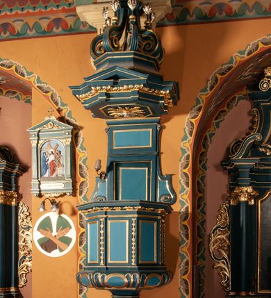 Zdjęcie nr 1: Ambona zawieszona na południowej ścianie pomiędzy ołtarzem św. Franciszka z Asyżu, a ołtarzem św. Kingi, dostępna schodami z grubości muru. Kosz na planie ośmioboku, z wypukłą dolną częścią podstawy zdobionej owalnymi, pustymi medalionami ujętymi uproszczonym puklowaniem. Ścianki kosza zdobione prostokątnymi płycinami, flankowanymi pilastrami o trzonach wypełnionych rzędem kampanul. Parapet kosza w formie prostego, profilowanego gzymsu. Zaplecek w kształcie stojącego prostokąta, dwudzielny, w każdej części dekorowany prostokątną płyciną, ujęty po bokach spływami wolutowymi. Baldachim na planie ośmioboku, z trzech stron z trójkątnymi przyczółkami, zaś belkowanie zdobione wicią liści akantu. W podniebiu baldachimu, w ośmiobocznej płycinie umieszczona gołębica Ducha Świętego na tle promienistej glorii. Zwieńczenie w kształcie sześciu wolut spiętych w kabłąk, przechodzących w prostopadłościenny szczyt z krzyżem łacińskim zatkniętym na obłokach. Każda z wolut zdobiona małym wazonikiem z listkami akantu. Struktura polichromowana na kolor ciemnozielony, detale i ornamentyka złocone i srebrzone. 

