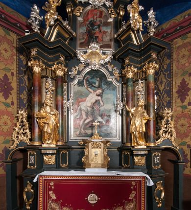 Zdjęcie nr 1: Nastawa architektoniczna, jednoosiowa z bramkami, jednokondygnacyjna ze zwieńczeniem, na dwustrefowym cokole. Ołtarz drewniany prostopadłościenny z kamienną mensą. Antepedium ujęte profilowaną ramą, wykonane z czerwonego, haftowanego aksamitu, w centrum medalion z przedstawieniem baranka leżącego na księdze, w dolnych narożach motywy kwiatowe. 
Retabulum flankowane ustawionymi pod kątem na zewnątrz dwiema parami korynckich kolumn, umieszczonych na tle kanelowanych pilastrów. Kolumny ustawione na cokołach, podtrzymują wyłamane belkowanie, przerwane nad polem środkowym, oraz przerwany falisty przyczółek. Na belkowaniu stoi para wazonów, na przerwanym przyczółku siedzą dwa putta. Pomiędzy kolumnami na cokołach znajdują się figury św. Jana Kantego i św. Jana Nepomucena. W polu głównym obraz z przedstawieniem św. Sebastiana ujęty profilowaną ramą w formie stojącego prostokąta zamkniętego łukiem półkolistym nadwieszonym z uskokiem, zwieńczoną rokokowym kartuszem flankowanym kwiatowymi zwisami. Zwieńczenie w formie stojącego prostokąta ujęte pilastrami o trzonach przechodzących w spływy wolutowe, zamknięte rokokowym kartuszem. W polu przedstawienie św. Floriana w profilowanej ramie w formie stojącego prostokąta zamkniętego łukiem półkolistym nadwieszonym. Bramki na wysokości cokołu nastawy w formie arkad podtrzymywanych przez pilastry, zwieńczonych racaille'owymi szczytami. Dwustrefowy cokół dekorowany rokokowymi płycinami, rocaille'm, rozetami i kartuszami. Kanelowane pilastry nastawy ozdobione kampanulami oraz bogatymi, kwiatowymi zwisami. Nastawa ujęta małymi uszami utworzonymi z liści palmy. 
Struktura architektoniczna polichromowana w kolorze czarnym, trzony kolumn pokryte polichromią imitującą kolorową marmoryzację, dekoracja rzeźbiarska, ornamentalna oraz profile pozłacane i posrebrzane. 

