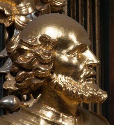 Zdjęcie nr 8: Rzeźba jest ścięta i wydrążona z tyłu, ustawiona na nieregularnej podstawie. Figura zwrócona jest frontalnie, w kontrapoście, z lekko pochylonymi głową i torsem, skierowanymi w lewo. Święty prawą ręką trzyma księgę opartą o jego biodro, lewą dłoń kładzie na piersi. Twarz ma wychudzoną, z podkreślonymi kośćmi policzkowymi, migdałowatymi, głęboko osadzonymi oczami, wąskim, małym nosem o ostrym profilu oraz wąskimi, otwartymi ustami, okoloną krótką, gęstą brodą, silnie wysuniętą do przodu oraz kręconymi włosami po bokach i z tyłu głowy. Święty ubrany jest w długą silnie fałdowaną tunikę oraz togę rektorską o szerokich rękawach, rozchyloną z przodu postaci z zygzakowato układającą się fałdą na jego prawej nodze. Figura w całości pozłocona. 
