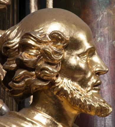 Zdjęcie nr 5: Rzeźba jest ścięta i wydrążona z tyłu, ustawiona na nieregularnej podstawie. Figura zwrócona jest frontalnie, w kontrapoście, z lekko pochylonymi głową i torsem, skierowanymi w lewo. Święty prawą ręką trzyma księgę opartą o jego biodro, lewą dłoń kładzie na piersi. Twarz ma wychudzoną, z podkreślonymi kośćmi policzkowymi, migdałowatymi, głęboko osadzonymi oczami, wąskim, małym nosem o ostrym profilu oraz wąskimi, otwartymi ustami, okoloną krótką, gęstą brodą, silnie wysuniętą do przodu oraz kręconymi włosami po bokach i z tyłu głowy. Święty ubrany jest w długą silnie fałdowaną tunikę oraz togę rektorską o szerokich rękawach, rozchyloną z przodu postaci z zygzakowato układającą się fałdą na jego prawej nodze. Figura w całości pozłocona. 
