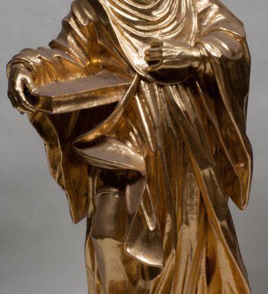 Zdjęcie nr 12: Rzeźba jest ścięta i wydrążona z tyłu, ustawiona na nieregularnej podstawie. Figura zwrócona jest frontalnie, w kontrapoście, z lekko pochylonymi głową i torsem, skierowanymi w lewo. Święty prawą ręką trzyma księgę opartą o jego biodro, lewą dłoń kładzie na piersi. Twarz ma wychudzoną, z podkreślonymi kośćmi policzkowymi, migdałowatymi, głęboko osadzonymi oczami, wąskim, małym nosem o ostrym profilu oraz wąskimi, otwartymi ustami, okoloną krótką, gęstą brodą, silnie wysuniętą do przodu oraz kręconymi włosami po bokach i z tyłu głowy. Święty ubrany jest w długą silnie fałdowaną tunikę oraz togę rektorską o szerokich rękawach, rozchyloną z przodu postaci z zygzakowato układającą się fałdą na jego prawej nodze. Figura w całości pozłocona. 
