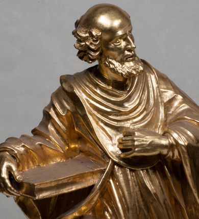 Zdjęcie nr 13: Rzeźba jest ścięta i wydrążona z tyłu, ustawiona na nieregularnej podstawie. Figura zwrócona jest frontalnie, w kontrapoście, z lekko pochylonymi głową i torsem, skierowanymi w lewo. Święty prawą ręką trzyma księgę opartą o jego biodro, lewą dłoń kładzie na piersi. Twarz ma wychudzoną, z podkreślonymi kośćmi policzkowymi, migdałowatymi, głęboko osadzonymi oczami, wąskim, małym nosem o ostrym profilu oraz wąskimi, otwartymi ustami, okoloną krótką, gęstą brodą, silnie wysuniętą do przodu oraz kręconymi włosami po bokach i z tyłu głowy. Święty ubrany jest w długą silnie fałdowaną tunikę oraz togę rektorską o szerokich rękawach, rozchyloną z przodu postaci z zygzakowato układającą się fałdą na jego prawej nodze. Figura w całości pozłocona. 
