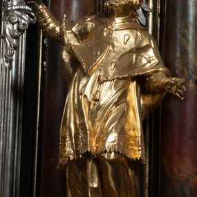 Zdjęcie nr 1: Rzeźba jest ścięta z tyłu i drążona, ustawiona na nieregularnej podstawie. Figura zwrócona jest frontalnie, w kontrapoście, z odchylonym w tył torsem i uniesioną głową, skierowaną w prawo. W prawej uniesionej wysoko ręce trzyma krucyfiks, lewą opuszczoną, wyciąga w bok. Twarz świętego jest szczupła, z uwypuklonymi kośćmi policzkowymi, migdałowatymi, głęboko osadzonymi oczami, wąskim, małym nosie o ostrym profilu oraz rozchylonymi, wąskimi ustami; okolona jest krótką, gęstą brodą wysuniętą do przodu oraz kręconymi, krótkimi włosami. Jan Nepomucen jest ubrany w długą sutannę, komżę dekorowaną przy dolnej krawędzi płaskorzeźbionym rocaille'm oraz w zawiązaną na piersi almucję. Figura jest w całości pozłocona. 






