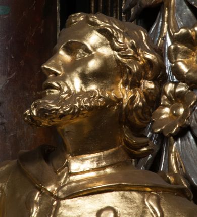Zdjęcie nr 5: Rzeźba jest ścięta z tyłu i drążona, ustawiona na nieregularnej podstawie. Figura zwrócona jest frontalnie, w kontrapoście, z odchylonym w tył torsem i uniesioną głową, skierowaną w prawo. W prawej uniesionej wysoko ręce trzyma krucyfiks, lewą opuszczoną, wyciąga w bok. Twarz świętego jest szczupła, z uwypuklonymi kośćmi policzkowymi, migdałowatymi, głęboko osadzonymi oczami, wąskim, małym nosie o ostrym profilu oraz rozchylonymi, wąskimi ustami; okolona jest krótką, gęstą brodą wysuniętą do przodu oraz kręconymi, krótkimi włosami. Jan Nepomucen jest ubrany w długą sutannę, komżę dekorowaną przy dolnej krawędzi płaskorzeźbionym rocaille&#039;m oraz w zawiązaną na piersi almucję. Figura jest w całości pozłocona. 






