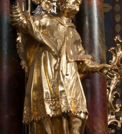 Zdjęcie nr 2: Rzeźba jest ścięta z tyłu i drążona, ustawiona na nieregularnej podstawie. Figura zwrócona jest frontalnie, w kontrapoście, z odchylonym w tył torsem i uniesioną głową, skierowaną w prawo. W prawej uniesionej wysoko ręce trzyma krucyfiks, lewą opuszczoną, wyciąga w bok. Twarz świętego jest szczupła, z uwypuklonymi kośćmi policzkowymi, migdałowatymi, głęboko osadzonymi oczami, wąskim, małym nosie o ostrym profilu oraz rozchylonymi, wąskimi ustami; okolona jest krótką, gęstą brodą wysuniętą do przodu oraz kręconymi, krótkimi włosami. Jan Nepomucen jest ubrany w długą sutannę, komżę dekorowaną przy dolnej krawędzi płaskorzeźbionym rocaille&#039;m oraz w zawiązaną na piersi almucję. Figura jest w całości pozłocona. 







