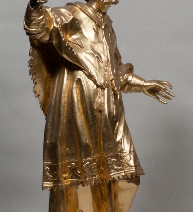 Zdjęcie nr 20: Rzeźba jest ścięta z tyłu i drążona, ustawiona na nieregularnej podstawie. Figura zwrócona jest frontalnie, w kontrapoście, z odchylonym w tył torsem i uniesioną głową, skierowaną w prawo. W prawej uniesionej wysoko ręce trzyma krucyfiks, lewą opuszczoną, wyciąga w bok. Twarz świętego jest szczupła, z uwypuklonymi kośćmi policzkowymi, migdałowatymi, głęboko osadzonymi oczami, wąskim, małym nosie o ostrym profilu oraz rozchylonymi, wąskimi ustami; okolona jest krótką, gęstą brodą wysuniętą do przodu oraz kręconymi, krótkimi włosami. Jan Nepomucen jest ubrany w długą sutannę, komżę dekorowaną przy dolnej krawędzi płaskorzeźbionym rocaille&#039;m oraz w zawiązaną na piersi almucję. Figura jest w całości pozłocona. 






