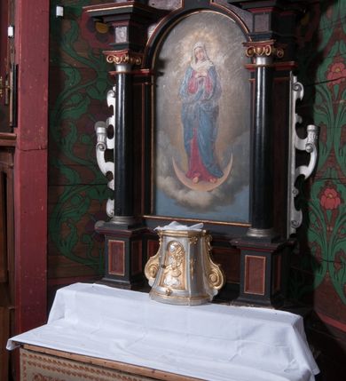 Zdjęcie nr 2: Nastawa ołtarzowa architektoniczna, jednokondygnacyjna, jednoosiowa na cokole. Ołtarz drewniany, prostopadłościenny, mensa drewniana. Antepedium w formie haftowanej maszynowo tkaniny. Nastawa ujęta parą pilastrów i parą stojących przed nimi kolumn o jońskich kapitelach dźwigających imposty o silnie wysuniętym gzymsie, na którym stoją dwa cokoły z figurami św. Biskupa i św. Floriana. Pole główne w całości wypełnione półkolistą arkadą podtrzymywaną na węgarach, zakończoną fragmentem gzymsu z uskrzydloną główką anielską. W polu obraz w formie stojącego prostokąta zamkniętego półkoliście z przedstawieniem Najświętszej Marii Panny Niepokalanie Poczętej. Kondygnacja zakończona fragmentem gzymsu, na którym stoi ośmioboczny cokół ujęty w narożach wolutami z koroną zamkniętą, zakończoną krzyżem. Nastawa ujęta uszami utworzonymi z ornamentu okuciowego. Struktura architektoniczna polichromowana na czarno, płyciny w kolorze czerwono-brązowym, profile pozłocone, uszy posrebrzone. 

