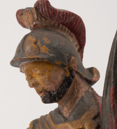 Zdjęcie nr 10: Figurka pełnoplastyczna, ustawiona na niewysokim, profilowanym cokole na planie prostokąta. Święty przedstawiony w całej postaci, zwrócony jest frontalnie, w wykroku, lekko pochylony w prawą stronę. Lewą ręką przytrzymuje przy boku chorągiew, prawą trzyma cebrzyk, z którego wylewa wodę na palący się budynek. Twarz ma szczupłą, okoloną krótką brodą. Ubrany jest w strój żołnierza: tunikę sięgającą kolan, zbroję torsową, na plecy ma zarzucony płaszcz, na głowie ma hełm z pióropuszem. Przy jego prawej nodze znajduje się niewielki budynek w płomieniach. Polichromia ciała naturalistyczna, broda brązowa, elementy stroju i chorągiew szaro-niebieskie z żółtymi detalami, płaszcz i pióropusz czerwone. 


