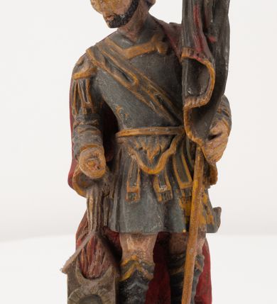 Zdjęcie nr 4: Figurka pełnoplastyczna, ustawiona na niewysokim, profilowanym cokole na planie prostokąta. Święty przedstawiony w całej postaci, zwrócony jest frontalnie, w wykroku, lekko pochylony w prawą stronę. Lewą ręką przytrzymuje przy boku chorągiew, prawą trzyma cebrzyk, z którego wylewa wodę na palący się budynek. Twarz ma szczupłą, okoloną krótką brodą. Ubrany jest w strój żołnierza: tunikę sięgającą kolan, zbroję torsową, na plecy ma zarzucony płaszcz, na głowie ma hełm z pióropuszem. Przy jego prawej nodze znajduje się niewielki budynek w płomieniach. Polichromia ciała naturalistyczna, broda brązowa, elementy stroju i chorągiew szaro-niebieskie z żółtymi detalami, płaszcz i pióropusz czerwone. 


