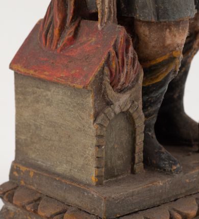 Zdjęcie nr 12: Figurka pełnoplastyczna, ustawiona na niewysokim, profilowanym cokole na planie prostokąta. Święty przedstawiony w całej postaci, zwrócony jest frontalnie, w wykroku, lekko pochylony w prawą stronę. Lewą ręką przytrzymuje przy boku chorągiew, prawą trzyma cebrzyk, z którego wylewa wodę na palący się budynek. Twarz ma szczupłą, okoloną krótką brodą. Ubrany jest w strój żołnierza: tunikę sięgającą kolan, zbroję torsową, na plecy ma zarzucony płaszcz, na głowie ma hełm z pióropuszem. Przy jego prawej nodze znajduje się niewielki budynek w płomieniach. Polichromia ciała naturalistyczna, broda brązowa, elementy stroju i chorągiew szaro-niebieskie z żółtymi detalami, płaszcz i pióropusz czerwone. 


