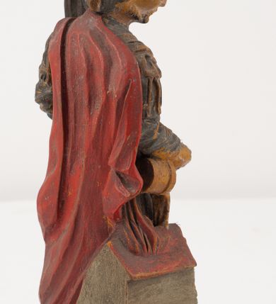 Zdjęcie nr 9: Figurka pełnoplastyczna, ustawiona na niewysokim, profilowanym cokole na planie prostokąta. Święty przedstawiony w całej postaci, zwrócony jest frontalnie, w wykroku, lekko pochylony w prawą stronę. Lewą ręką przytrzymuje przy boku chorągiew, prawą trzyma cebrzyk, z którego wylewa wodę na palący się budynek. Twarz ma szczupłą, okoloną krótką brodą. Ubrany jest w strój żołnierza: tunikę sięgającą kolan, zbroję torsową, na plecy ma zarzucony płaszcz, na głowie ma hełm z pióropuszem. Przy jego prawej nodze znajduje się niewielki budynek w płomieniach. Polichromia ciała naturalistyczna, broda brązowa, elementy stroju i chorągiew szaro-niebieskie z żółtymi detalami, płaszcz i pióropusz czerwone. 


