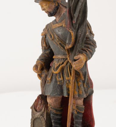 Zdjęcie nr 1: Figurka pełnoplastyczna, ustawiona na niewysokim, profilowanym cokole na planie prostokąta. Święty przedstawiony w całej postaci, zwrócony jest frontalnie, w wykroku, lekko pochylony w prawą stronę. Lewą ręką przytrzymuje przy boku chorągiew, prawą trzyma cebrzyk, z którego wylewa wodę na palący się budynek. Twarz ma szczupłą, okoloną krótką brodą. Ubrany jest w strój żołnierza: tunikę sięgającą kolan, zbroję torsową, na plecy ma zarzucony płaszcz, na głowie ma hełm z pióropuszem. Przy jego prawej nodze znajduje się niewielki budynek w płomieniach. Polichromia ciała naturalistyczna, broda brązowa, elementy stroju i chorągiew szaro-niebieskie z żółtymi detalami, płaszcz i pióropusz czerwone. 


