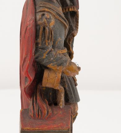 Zdjęcie nr 6: Figurka pełnoplastyczna, ustawiona na niewysokim, profilowanym cokole na planie prostokąta. Święty przedstawiony w całej postaci, zwrócony jest frontalnie, w wykroku, lekko pochylony w prawą stronę. Lewą ręką przytrzymuje przy boku chorągiew, prawą trzyma cebrzyk, z którego wylewa wodę na palący się budynek. Twarz ma szczupłą, okoloną krótką brodą. Ubrany jest w strój żołnierza: tunikę sięgającą kolan, zbroję torsową, na plecy ma zarzucony płaszcz, na głowie ma hełm z pióropuszem. Przy jego prawej nodze znajduje się niewielki budynek w płomieniach. Polichromia ciała naturalistyczna, broda brązowa, elementy stroju i chorągiew szaro-niebieskie z żółtymi detalami, płaszcz i pióropusz czerwone. 



