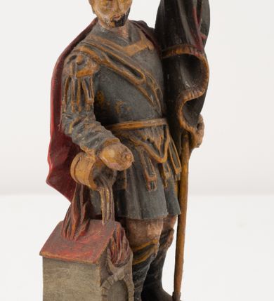 Zdjęcie nr 5: Figurka pełnoplastyczna, ustawiona na niewysokim, profilowanym cokole na planie prostokąta. Święty przedstawiony w całej postaci, zwrócony jest frontalnie, w wykroku, lekko pochylony w prawą stronę. Lewą ręką przytrzymuje przy boku chorągiew, prawą trzyma cebrzyk, z którego wylewa wodę na palący się budynek. Twarz ma szczupłą, okoloną krótką brodą. Ubrany jest w strój żołnierza: tunikę sięgającą kolan, zbroję torsową, na plecy ma zarzucony płaszcz, na głowie ma hełm z pióropuszem. Przy jego prawej nodze znajduje się niewielki budynek w płomieniach. Polichromia ciała naturalistyczna, broda brązowa, elementy stroju i chorągiew szaro-niebieskie z żółtymi detalami, płaszcz i pióropusz czerwone. 


