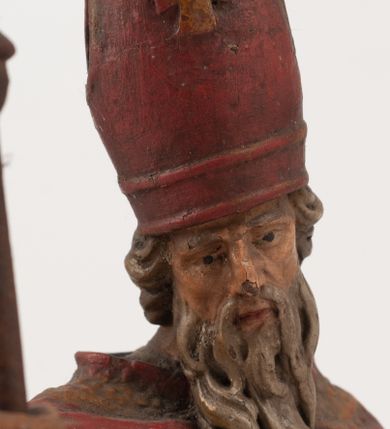 Zdjęcie nr 11: Figura jest pełnoplastyczna, ustawiona na profilowanym cokole na planie prostokąta. Święty przedstawiony w całej postaci, zwrócony jest frontalnie, w kontrapoście, z głową skierowaną w prawo. Obie ręce wyciąga przed siebie, w prawej trzyma pastorał. Twarz ma pociągłą, o  oczach skierowanych w dół, dużym, garbatym nosie i niewielkich ustach, okoloną długą brodą uformowaną w pojedyncze pukle, opadającą na pierś oraz krótkimi, silnie kręconymi włosami. Święty ubrany jest w strój biskupa: białą albę przepasaną w talii, czerwoną stułę, czerwoną kapę z niebieską podszewką, zapiętą na piersi, na głowie ma czerwoną infułę. Polichromia ciała naturalistyczna. 
