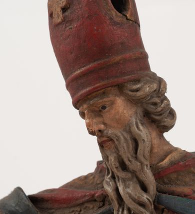 Zdjęcie nr 12: Figura jest pełnoplastyczna, ustawiona na profilowanym cokole na planie prostokąta. Święty przedstawiony w całej postaci, zwrócony jest frontalnie, w kontrapoście, z głową skierowaną w prawo. Obie ręce wyciąga przed siebie, w prawej trzyma pastorał. Twarz ma pociągłą, o  oczach skierowanych w dół, dużym, garbatym nosie i niewielkich ustach, okoloną długą brodą uformowaną w pojedyncze pukle, opadającą na pierś oraz krótkimi, silnie kręconymi włosami. Święty ubrany jest w strój biskupa: białą albę przepasaną w talii, czerwoną stułę, czerwoną kapę z niebieską podszewką, zapiętą na piersi, na głowie ma czerwoną infułę. Polichromia ciała naturalistyczna. 
