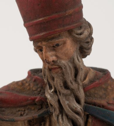 Zdjęcie nr 13: Figura jest pełnoplastyczna, ustawiona na profilowanym cokole na planie prostokąta. Święty przedstawiony w całej postaci, zwrócony jest frontalnie, w kontrapoście, z głową skierowaną w prawo. Obie ręce wyciąga przed siebie, w prawej trzyma pastorał. Twarz ma pociągłą, o  oczach skierowanych w dół, dużym, garbatym nosie i niewielkich ustach, okoloną długą brodą uformowaną w pojedyncze pukle, opadającą na pierś oraz krótkimi, silnie kręconymi włosami. Święty ubrany jest w strój biskupa: białą albę przepasaną w talii, czerwoną stułę, czerwoną kapę z niebieską podszewką, zapiętą na piersi, na głowie ma czerwoną infułę. Polichromia ciała naturalistyczna. 
