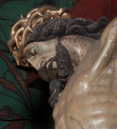 Zdjęcie nr 5: Figura umarłego Chrystusa przybita jest trzema gwoździami do prostego krzyża. Ciało w lekkim zwisie, o szeroko rozłożonych ramionach, układających się w delikatny łuk, głowa opada bezwładnie na prawe ramię, nogi są delikatnie ugięte w kolanach, stopy skrzyżowane w układzie prawa na lewą. Twarz pociągła, o łagodnych rysach i niskim czole, zamkniętych oczach, dużym nosie i rozchylonych ustach, okolona krótką, rozdwajającą się brodą oraz silnie kręconymi włosami opadającymi na prawy bark. Na głowie korona cierniowa. Ciało wychudzone, smukłe, o miękko modelowanej muskulaturze oraz silnie zaznaczonych żebrach i mostku klatki piersiowej, z podkreślonymi liniami ścięgien w partiach ramion i nóg. Biodra przepasane ekspresyjnie drapowanym perizonium zawieszonym na podwójnym sznurze, sięgającym połowy ud, odsłaniającym prawe biodro, ze zwisem rozwianej tkaniny na prawym boku. Polichromia naturalistyczna w partiach ciała z zaznaczonymi śladami męki, perizonium i korona cierniowa pozłacane. W górnej części krzyża banderola z inskrypcją „I.N. / R.I.”.



