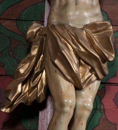 Zdjęcie nr 6: Figura umarłego Chrystusa przybita jest trzema gwoździami do prostego krzyża. Ciało w lekkim zwisie, o szeroko rozłożonych ramionach, układających się w delikatny łuk, głowa opada bezwładnie na prawe ramię, nogi są delikatnie ugięte w kolanach, stopy skrzyżowane w układzie prawa na lewą. Twarz pociągła, o łagodnych rysach i niskim czole, zamkniętych oczach, dużym nosie i rozchylonych ustach, okolona krótką, rozdwajającą się brodą oraz silnie kręconymi włosami opadającymi na prawy bark. Na głowie korona cierniowa. Ciało wychudzone, smukłe, o miękko modelowanej muskulaturze oraz silnie zaznaczonych żebrach i mostku klatki piersiowej, z podkreślonymi liniami ścięgien w partiach ramion i nóg. Biodra przepasane ekspresyjnie drapowanym perizonium zawieszonym na podwójnym sznurze, sięgającym połowy ud, odsłaniającym prawe biodro, ze zwisem rozwianej tkaniny na prawym boku. Polichromia naturalistyczna w partiach ciała z zaznaczonymi śladami męki, perizonium i korona cierniowa pozłacane. W górnej części krzyża banderola z inskrypcją „I.N. / R.I.”.



