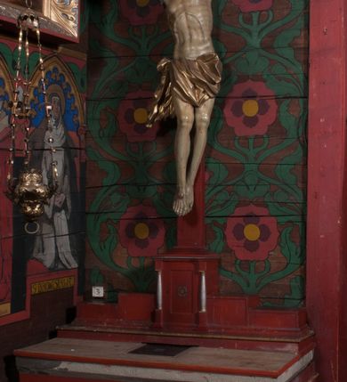 Zdjęcie nr 2: Figura umarłego Chrystusa przybita jest trzema gwoździami do prostego krzyża. Ciało w lekkim zwisie, o szeroko rozłożonych ramionach, układających się w delikatny łuk, głowa opada bezwładnie na prawe ramię, nogi są delikatnie ugięte w kolanach, stopy skrzyżowane w układzie prawa na lewą. Twarz pociągła, o łagodnych rysach i niskim czole, zamkniętych oczach, dużym nosie i rozchylonych ustach, okolona krótką, rozdwajającą się brodą oraz silnie kręconymi włosami opadającymi na prawy bark. Na głowie korona cierniowa. Ciało wychudzone, smukłe, o miękko modelowanej muskulaturze oraz silnie zaznaczonych żebrach i mostku klatki piersiowej, z podkreślonymi liniami ścięgien w partiach ramion i nóg. Biodra przepasane ekspresyjnie drapowanym perizonium zawieszonym na podwójnym sznurze, sięgającym połowy ud, odsłaniającym prawe biodro, ze zwisem rozwianej tkaniny na prawym boku. Polichromia naturalistyczna w partiach ciała z zaznaczonymi śladami męki, perizonium i korona cierniowa pozłacane. W górnej części krzyża banderola z inskrypcją „I.N. / R.I.”.



