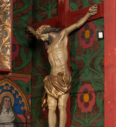 Zdjęcie nr 1: Figura umarłego Chrystusa przybita jest trzema gwoździami do prostego krzyża. Ciało w lekkim zwisie, o szeroko rozłożonych ramionach, układających się w delikatny łuk, głowa opada bezwładnie na prawe ramię, nogi są delikatnie ugięte w kolanach, stopy skrzyżowane w układzie prawa na lewą. Twarz pociągła, o łagodnych rysach i niskim czole, zamkniętych oczach, dużym nosie i rozchylonych ustach, okolona krótką, rozdwajającą się brodą oraz silnie kręconymi włosami opadającymi na prawy bark. Na głowie korona cierniowa. Ciało wychudzone, smukłe, o miękko modelowanej muskulaturze oraz silnie zaznaczonych żebrach i mostku klatki piersiowej, z podkreślonymi liniami ścięgien w partiach ramion i nóg. Biodra przepasane ekspresyjnie drapowanym perizonium zawieszonym na podwójnym sznurze, sięgającym połowy ud, odsłaniającym prawe biodro, ze zwisem rozwianej tkaniny na prawym boku. Polichromia naturalistyczna w partiach ciała z zaznaczonymi śladami męki, perizonium i korona cierniowa pozłacane. W górnej części krzyża banderola z inskrypcją „I.N. / R.I.”.



