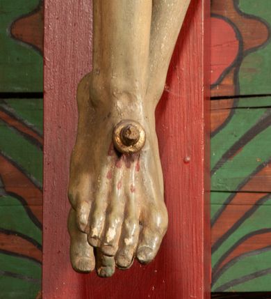 Zdjęcie nr 8: Figura umarłego Chrystusa przybita jest trzema gwoździami do prostego krzyża. Ciało w lekkim zwisie, o szeroko rozłożonych ramionach, układających się w delikatny łuk, głowa opada bezwładnie na prawe ramię, nogi są delikatnie ugięte w kolanach, stopy skrzyżowane w układzie prawa na lewą. Twarz pociągła, o łagodnych rysach i niskim czole, zamkniętych oczach, dużym nosie i rozchylonych ustach, okolona krótką, rozdwajającą się brodą oraz silnie kręconymi włosami opadającymi na prawy bark. Na głowie korona cierniowa. Ciało wychudzone, smukłe, o miękko modelowanej muskulaturze oraz silnie zaznaczonych żebrach i mostku klatki piersiowej, z podkreślonymi liniami ścięgien w partiach ramion i nóg. Biodra przepasane ekspresyjnie drapowanym perizonium zawieszonym na podwójnym sznurze, sięgającym połowy ud, odsłaniającym prawe biodro, ze zwisem rozwianej tkaniny na prawym boku. Polichromia naturalistyczna w partiach ciała z zaznaczonymi śladami męki, perizonium i korona cierniowa pozłacane. W górnej części krzyża banderola z inskrypcją „I.N. / R.I.”.



