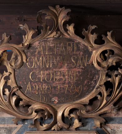 Zdjęcie nr 4: Zwieńczenie w formie stojącego prostokąta lekko rozszerzającego się u dołu, w jego narożach trójkątne płyciny. Flankowane jest uszami z suchego akantu wyrastającego z woluty (prawego brak). Zwieńczone jest owalnym kartuszem ujętym obramieniem z suchego akantu, z napisem „ALTARE / OMNIVM SAN/CTORVM / ANNO 1709/ Die [...] 8clo”. W polu środkowym obraz Wszystkich Świętych w sześciobocznej ramie dekorowanej suchym akantem i rozetkami. W centrum przedstawienia Matka Boska na obłoku w otoczeniu świętych. Zwrócona jest frontalnie, z dłońmi złożonymi w geście modlitwy i głową skierowaną do góry. Po jej prawej stronie można rozpoznać św. Piotra, który został ukazany jako starzec, trzymający w dłoni klucze. Po jej lewej stronie znajduje się św. Jan Chrzciciel, ubrany w melotę, trzymający laskę zakończoną krzyżem z banderolą. Na pierwszym planie, po dwóch stronach przedstawienia znajdują się rycerze w zbrojach, na koniach, zwróceni do wewnątrz. Rycerz po prawej stronie to św. Jerzy, który włócznią zabija smoka. Po lewej stronie znajduje się św. Michał Archanioł, którego koń stoi na tylnych kopytach, a przednimi tratuje szatana. W tle liczne zastępy świętych. Wszystkie postacie, oprócz rycerzy, z głowami zwróconymi ku górze, gdzie na tle obłoków przedstawione jest oko Opatrzności. Kolorystyka ciepła, utrzymana w tonacji żółto-brązowej z dodatkiem błękitu i czerwieni. Struktura zwieńczenia polichromowana w kolorze niebieskim i szarym, z pozłacaną ornamentyką i profilami. 
