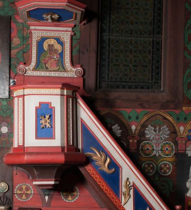 Zdjęcie nr 2: Ambona przyścienna, wisząca, z dostępem od wschodu w postaci jednobiegowych schodów z balustradą. Kosz na planie kwadratu o ściętych narożach, na podstawie w formie wałka, z podwieszeniem o wklęsłych bokach, zakończonym gzymsem. Zachodnia i południowa ścianka kosza ujęte pilastrami, parapet w formie profilowanego gzymsu. Ścianka frontowa ambony dekorowana płyciną w kształcie stojącego prostokąta o wciętych górnych narożach z rozetą w polu, zachodnia zaś liściem palmy. Balustrada schodów dekorowana dwoma płycinami z liśćmi palmowymi, poręcz w formie profilowanego gzymsu. Zaplecek w kształcie stojącego prostokąta ujętego spływami wolutowymi. W profilowanej płycinie o fantazyjnym wykroju malowane przedstawienie Matki Boskiej z Dzieciątkiem. Baldachim na planie kwadratu o ściętych narożach, zwieńczony wielobocznym postumentem, o wklęsłych ściankach, na którym znajduje się krzyż z czworoliściem w środku. Na skrzyżowaniu ramion krzyża wiązki promieni. Na podniebiu baldachimu, w ośmiobocznej płycinie gołębica Ducha Świętego w glorii promienistej. Struktura ambony polichromowana w kolorze białym, z czerwonymi cokołami, obramieniami i profilami, pola płycin niebieskie.  Pilastry kosza, pas balustrady, zaplecek i podniebie baldachimu dekorowane pozłoconym ornamentem geometrycznym.

