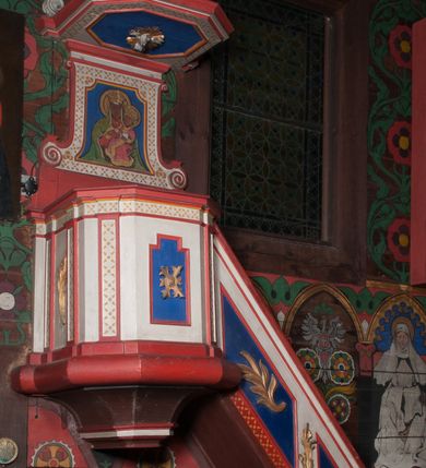 Zdjęcie nr 1: Ambona przyścienna, wisząca, z dostępem od wschodu w postaci jednobiegowych schodów z balustradą. Kosz na planie kwadratu o ściętych narożach, na podstawie w formie wałka, z podwieszeniem o wklęsłych bokach, zakończonym gzymsem. Zachodnia i południowa ścianka kosza ujęte pilastrami, parapet w formie profilowanego gzymsu. Ścianka frontowa ambony dekorowana płyciną w kształcie stojącego prostokąta o wciętych górnych narożach z rozetą w polu, zachodnia zaś liściem palmy. Balustrada schodów dekorowana dwoma płycinami z liśćmi palmowymi, poręcz w formie profilowanego gzymsu. Zaplecek w kształcie stojącego prostokąta ujętego spływami wolutowymi. W profilowanej płycinie o fantazyjnym wykroju malowane przedstawienie Matki Boskiej z Dzieciątkiem. Baldachim na planie kwadratu o ściętych narożach, zwieńczony wielobocznym postumentem, o wklęsłych ściankach, na którym znajduje się krzyż z czworoliściem w środku. Na skrzyżowaniu ramion krzyża wiązki promieni. Na podniebiu baldachimu, w ośmiobocznej płycinie gołębica Ducha Świętego w glorii promienistej. Struktura ambony polichromowana w kolorze białym, z czerwonymi cokołami, obramieniami i profilami, pola płycin niebieskie.  Pilastry kosza, pas balustrady, zaplecek i podniebie baldachimu dekorowane pozłoconym ornamentem geometrycznym.
