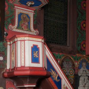 Zdjęcie nr 1: Ambona przyścienna, wisząca, z dostępem od wschodu w postaci jednobiegowych schodów z balustradą. Kosz na planie kwadratu o ściętych narożach, na podstawie w formie wałka, z podwieszeniem o wklęsłych bokach, zakończonym gzymsem. Zachodnia i południowa ścianka kosza ujęte pilastrami, parapet w formie profilowanego gzymsu. Ścianka frontowa ambony dekorowana płyciną w kształcie stojącego prostokąta o wciętych górnych narożach z rozetą w polu, zachodnia zaś liściem palmy. Balustrada schodów dekorowana dwoma płycinami z liśćmi palmowymi, poręcz w formie profilowanego gzymsu. Zaplecek w kształcie stojącego prostokąta ujętego spływami wolutowymi. W profilowanej płycinie o fantazyjnym wykroju malowane przedstawienie Matki Boskiej z Dzieciątkiem. Baldachim na planie kwadratu o ściętych narożach, zwieńczony wielobocznym postumentem, o wklęsłych ściankach, na którym znajduje się krzyż z czworoliściem w środku. Na skrzyżowaniu ramion krzyża wiązki promieni. Na podniebiu baldachimu, w ośmiobocznej płycinie gołębica Ducha Świętego w glorii promienistej. Struktura ambony polichromowana w kolorze białym, z czerwonymi cokołami, obramieniami i profilami, pola płycin niebieskie.  Pilastry kosza, pas balustrady, zaplecek i podniebie baldachimu dekorowane pozłoconym ornamentem geometrycznym.
