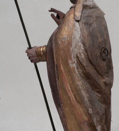 Zdjęcie nr 2: Figura nieopracowana rzeźbiarsko z tyłu, ustawiona jest na niewielkiej, nieregularnej podstawie. Święty ukazany jest w całej postaci, frontalnie, z głową skierowaną w prawo. Prawą rękę unosi delikatnie w geście błogosławieństwa, w lewej wyciągniętej do przodu trzyma laskę zakończoną krzyżem. Jego twarz jest trójkątna, o półprzymkniętych oczach, niewielkim nosie i wąskich ustach. Ubrany jest w albę, ornat z płaskorzeźbioną krzyżową pretekstą, kapę zapiętą na piersi, na głowie ma infułę. Przy jego prawej nodze znajduje się półpostać Piotrowina, zwróconego ku świętemu,  z rękoma złożonymi w geście modlitwy, nakrytego płaszczem. Polichromia w odsłoniętych partiach karnacji obecnie poszarzała, strój złocony. 







