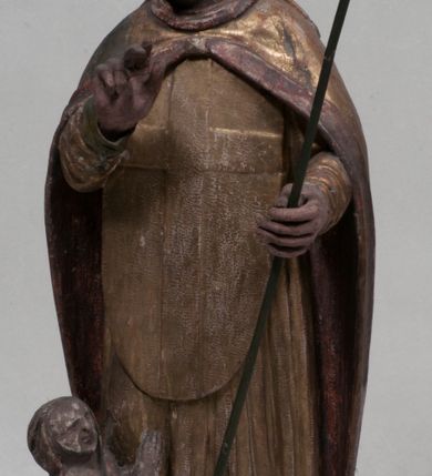 Zdjęcie nr 1: Figura nieopracowana rzeźbiarsko z tyłu, ustawiona jest na niewielkiej, nieregularnej podstawie. Święty ukazany jest w całej postaci, frontalnie, z głową skierowaną w prawo. Prawą rękę unosi delikatnie w geście błogosławieństwa, w lewej wyciągniętej do przodu trzyma laskę zakończoną krzyżem. Jego twarz jest trójkątna, o półprzymkniętych oczach, niewielkim nosie i wąskich ustach. Ubrany jest w albę, ornat z płaskorzeźbioną krzyżową pretekstą, kapę zapiętą na piersi, na głowie ma infułę. Przy jego prawej nodze znajduje się półpostać Piotrowina, zwróconego ku świętemu,  z rękoma złożonymi w geście modlitwy, nakrytego płaszczem. Polichromia w odsłoniętych partiach karnacji obecnie poszarzała, strój złocony. 







